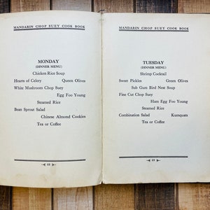 Pu&ograve; includere: Libro di cucina aperto, intitolato "Mandarin Chop Suey Cook Book", che mostra due pagine affiancate. La pagina di sinistra elenca un menu per la cena del luned&igrave;, tra cui Zuppa di pollo e Egg Foo Young. La pagina di destra elenca un menu per la cena del marted&igrave; con Cocktail di gamberetti e Kumquat.