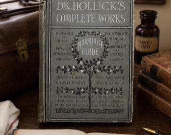 May include: An antique book titled "Dr. Hollick's Complete Works" with the text "Marriage Guide" in a floral wreath. The book is open to a page with the text "Origin of Life". Other books and a framed sign are in the background.