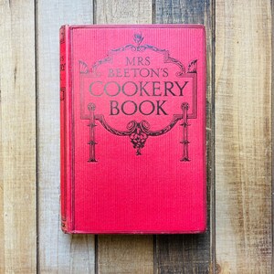 Pode incluir: Um livro vermelho de capa dura com o t&iacute;tulo "MRS. BEETON'S COOKERY BOOK" em letras pretas.