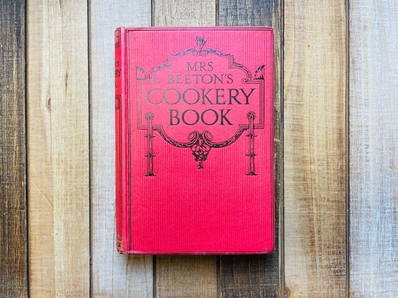 Pode incluir: Um livro vermelho de capa dura com o t&iacute;tulo "MRS. BEETON'S COOKERY BOOK" em letras pretas.