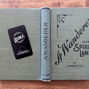 May include: An open, vintage book titled "A Wanderer in the Spirit Lands" by Franchezzo. The book has a light green cover with black lettering and decorative elements. A black business card from Jerry's Ephemera rests on the left side.
