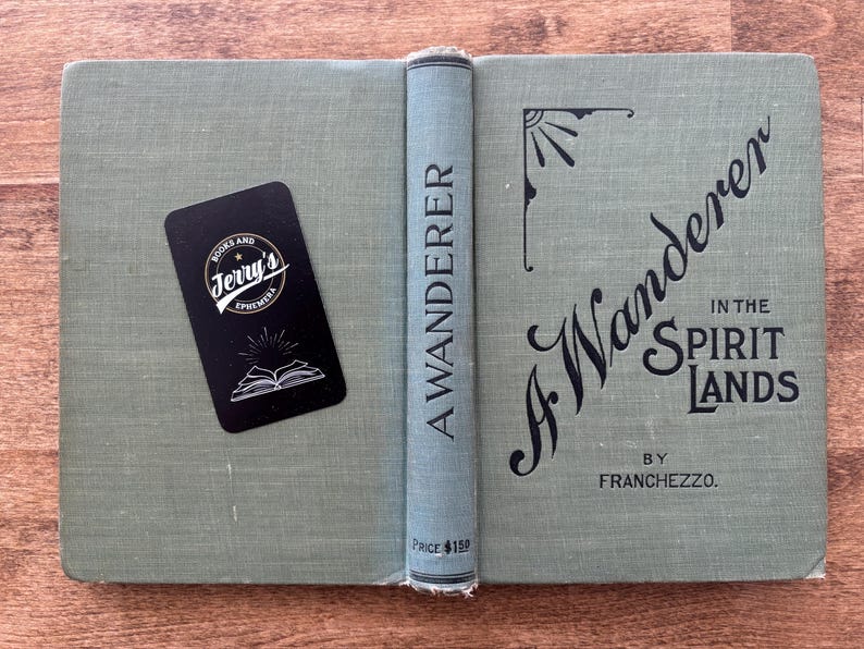 May include: An open, vintage book titled "A Wanderer in the Spirit Lands" by Franchezzo. The book has a light green cover with black lettering and decorative elements. A black business card from Jerry's Ephemera rests on the left side.