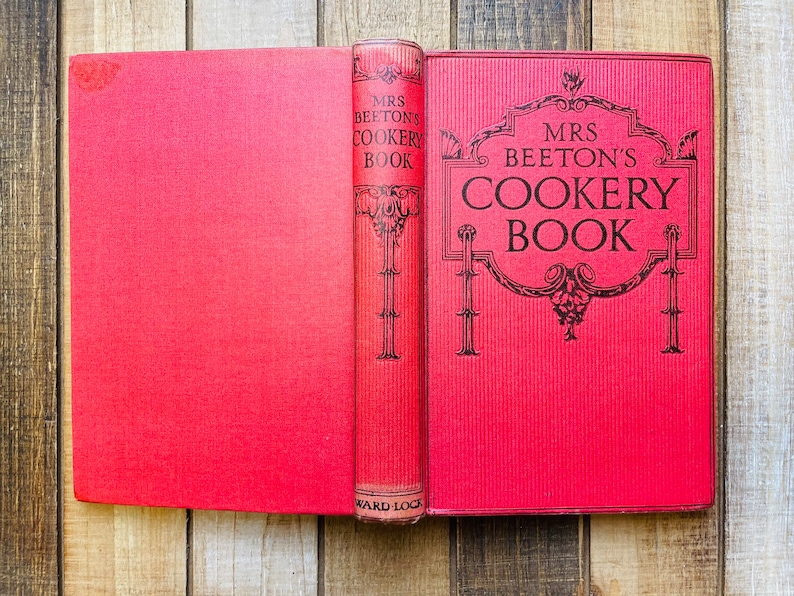 Pode incluir: Um livro vermelho de capa dura intitulado "Mrs. Beeton's Cookery Book" com um design decorativo dourado na capa. O livro est&aacute; aberto para revelar uma p&aacute;gina em branco.