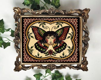 May include: A framed art print featuring a woman with butterfly wings. The artwork has a vintage tattoo style, with a cream background and a decorative gold frame. The butterfly wings are red, green, and black.