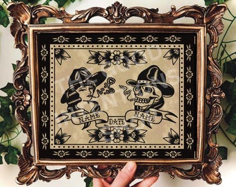 May include: A framed tattoo-style print featuring a woman and a skull, both wearing cowboy hats and smoking. The black and tan artwork includes banners with the words "NAME" and "DATE". The ornate frame is bronze-colored.