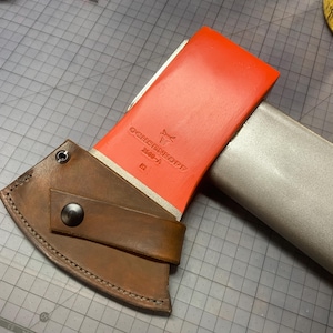 May include: A red axe head with the text "GOREN KOPP" and a brown leather sheath. The axe has a wooden handle and a gray protective cover. The sheath is secured with a black snap.
