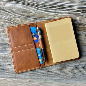 May include: A brown leather notebook cover with a pen, credit cards, and a small notepad. The cover has a stitched design and elastic band. The notepad is open, revealing blank pages. The pen is silver and black.