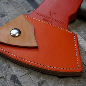May include: Orange leather axe sheath with a tan leather flap and a silver snap closure. The sheath has white stitching and the text "ÖCHSNERKÖFE 1200-A" is embossed on the front.