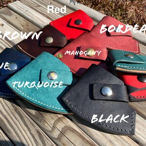 Pu&ograve; includere: Una collezione di portafogli in pelle fatti a mano in vari colori, tra cui rosso, marrone, blu, turchese e nero. Ogni portafoglio &egrave; dotato di chiusura a scatto ed &egrave; esposto su una superficie di legno. I colori sono etichettati con testo bianco.