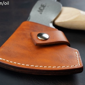 Pu&ograve; includere: Primo piano di un fodero per ascia in pelle marrone sella con cuciture bianche e una chiusura a scatto argentata. La testa dell'ascia &egrave; parzialmente visibile, insieme a un manico in legno. Il testo "Saddle tan/oil" &egrave; in alto a sinistra.