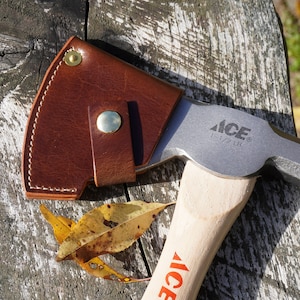 May include: A hatchet with a wooden handle and a metal head, partially covered by a brown leather sheath. The head is stamped with "ACE" and "1-1/2 LB". The hatchet rests on a weathered wooden surface, with a few fallen leaves.