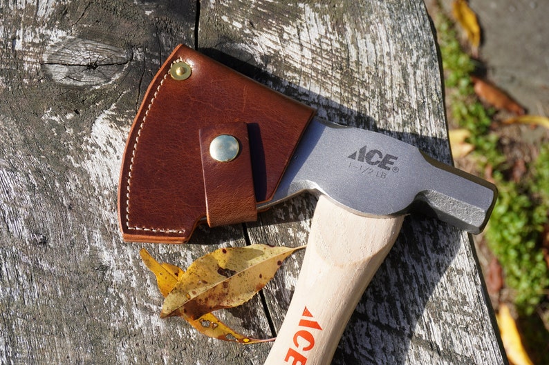 Pu&ograve; includere: Un'ascia con manico in legno e testa in metallo, parzialmente coperta da un fodero in pelle marrone. La testa &egrave; stampata con "ACE" e "1-1/2 LB". L'ascia poggia su una superficie di legno stagionato, con alcune foglie cadute.