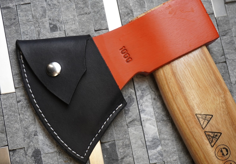 May include: A black leather axe sheath with a silver button and white stitching. The sheath is attached to a wooden-handled axe with an orange head. The axe head has the number "1000" printed on it.