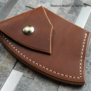 May include: A brown leather knife sheath with a star-shaped snap closure. The sheath has a stitched edge and is designed to hold a small knife.