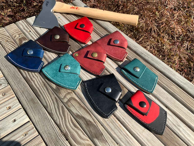 Pu&ograve; includere: Una collezione di fodere per ascia in pelle di vari colori, tra cui rosso, blu e nero, con chiusure a scatto argentate. Un'ascia con manico in legno e il logo "ACE" &egrave; visibile sullo sfondo. Le fodere sono disposte su una superficie di legno.