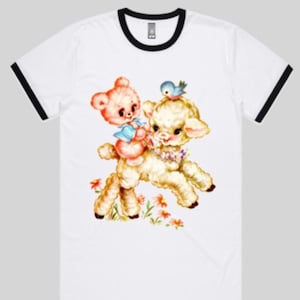 May include: A white t-shirt with a black ringer neckline features a vintage illustration of a pink teddy bear riding a white lamb with a blue bird perched on the lamb's head.