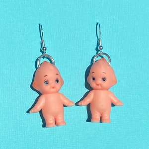 May include: A pair of silver dangle earrings featuring small pink plastic dolls with blue eyes. The dolls are wearing no clothing and have a simple, sweet expression.