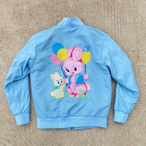 May include: A light blue bomber jacket with an embroidered design of a pink bunny holding balloons and a smaller yellow bunny. The balloons are yellow, pink, and blue.