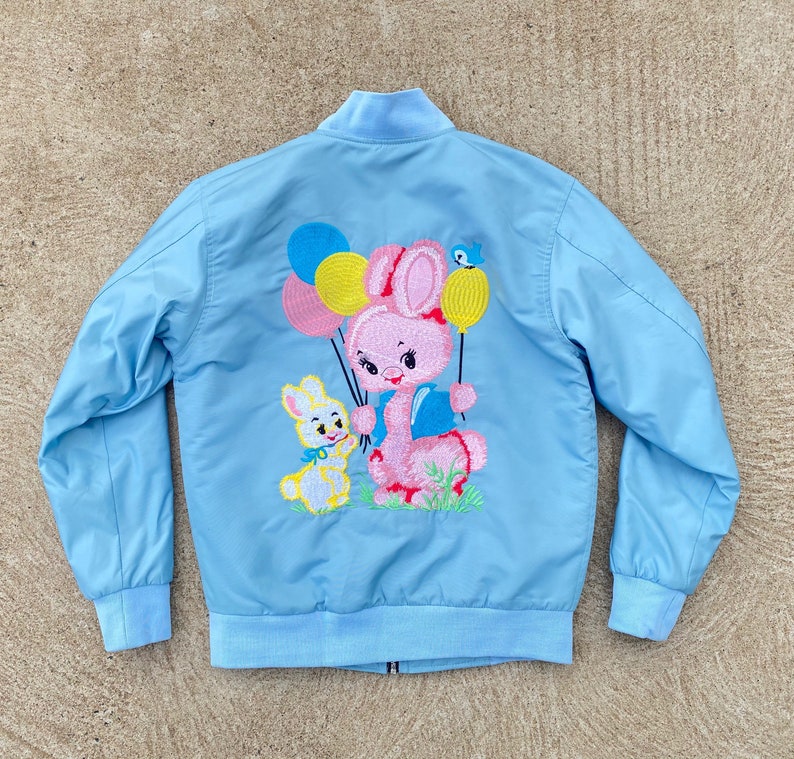 May include: A light blue bomber jacket with an embroidered design of a pink bunny holding balloons and a smaller yellow bunny. The balloons are yellow, pink, and blue.