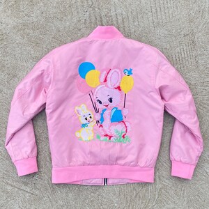 May include: A pink bomber jacket with an embroidered design of a pink bunny holding balloons and a smaller yellow bunny. The balloons are yellow, blue, and pink.