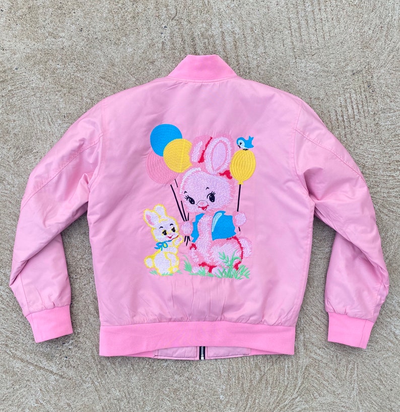 May include: A pink bomber jacket with an embroidered design of a pink bunny holding balloons and a smaller yellow bunny. The balloons are yellow, blue, and pink.