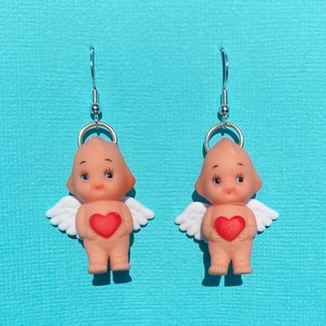 May include: A pair of dangle earrings featuring a small plastic cherub holding a red heart. The cherubs have white wings and are wearing a halo. The earrings are silver.