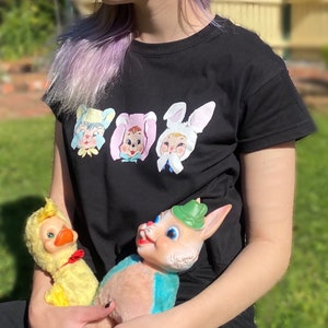 Puede incluir: Una camiseta negra con un estampado gráfico de tres caras de conejitos de dibujos animados. La persona está sosteniendo dos peluches, un pato amarillo y un conejito rosa y azul.