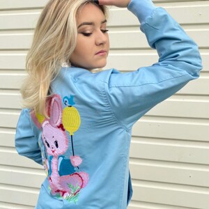 May include: A light blue bomber jacket with a pink embroidered bunny holding a yellow balloon. The bunny is wearing a blue shirt and has a pink nose.