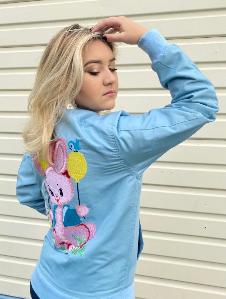 May include: A light blue bomber jacket with a pink embroidered bunny holding a yellow balloon. The bunny is wearing a blue shirt and has a pink nose.