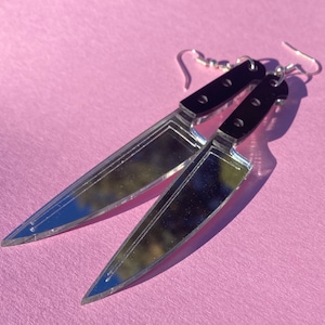 May include: A pair of silver knife earrings with black plastic handles. The earrings are dangling and have a reflective surface.