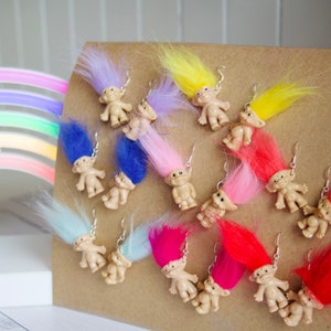 Op de afbeelding: Een verzameling van 12 paar trollenpop oorbellen. Elk paar heeft een kleine plastic trollenpop met kleurrijk, pluche haar. De trollen zijn gerangschikt op een bruine achtergrond.