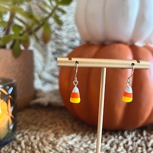 May include: A pair of silver dangle earrings with candy corn charms. The charms are orange, white, and yellow.