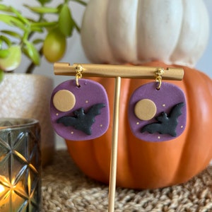 Peut inclure: Une paire de boucles d'oreilles carrées violettes avec une lune dorée et une silhouette de chauve-souris noire. Les boucles d'oreilles sont sur un support doré.