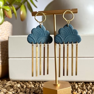 May include: A pair of blue cloud-shaped earrings with gold dangling chains. The earrings are displayed on a gold stand.