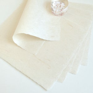 May include: Off-white handmade paper sheets with deckled edges, stacked and partially rolled. A small, clear glass votive candle holder with a floral design sits on top of the paper. The paper has a textured, fibrous appearance.