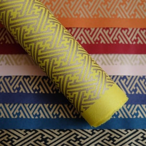 May include: A roll of yellow wrapping paper with a geometric pattern. The paper is rolled up and the end is visible. The paper is on top of a stack of colorful patterned paper.