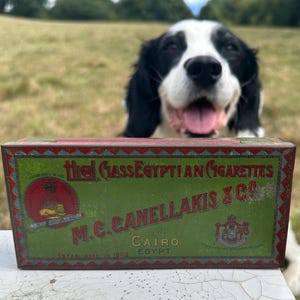 May include: An antique green and red cigarette tin with the text "High Class Egyptian Cigarettes" and "M.C. Canellakis & Co. Cairo Egypt". A black and white dog is in the background.