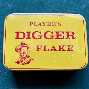 May include: A rectangular yellow tin with red text and a red border. The text reads "PLAYER'S DIGGER FLAKE" with an illustration of a man in a hat smoking a pipe. The tin has a slightly speckled appearance.