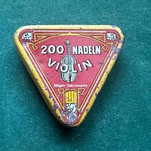 Puede incluir: Una lata triangular vintage con centro rojo y borde amarillo. La lata dice "200 NADELN VIOLIN" con una ilustración de violín. La lata tiene una apariencia desgastada, lo que sugiere edad y uso. El fondo es una superficie verde.