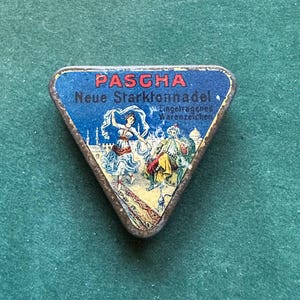 May include: A triangular vintage tin with a blue background, featuring the word "PASCHA" in red. The tin displays an illustration of a dancing woman and a man with a hookah. The text "Neue Starktonnadel" is also visible.
