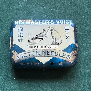 Op de afbeelding: Een rechthoekig vintage blikje van "VICTOR NEEDLES" met een blauw en wit ontwerp. Het blikje toont de zin "HIS MASTER'S VOICE" en een illustratie van een hond die naar een grammofoon luistert. Japanse karakters zijn ook aanwezig.