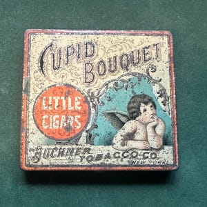 May include: Vintage square tin with the words "Cupid Bouquet" and "Little Cigars" in distressed lettering. Features an illustration of a cherub with wings and the text "Buchner Tobacco Co. New York". The tin has a worn, aged appearance.