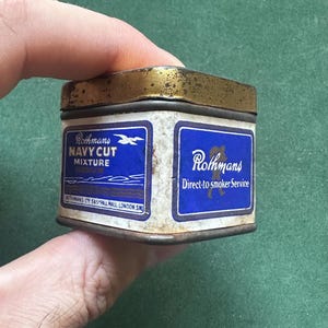 May include: A rectangular Rothmans Navy Cut Mixture tin with a gold-colored lid and blue and white labels. The labels display the Rothmans logo and text, including "Navy Cut Mixture" and "Direct-to-smoker Service."