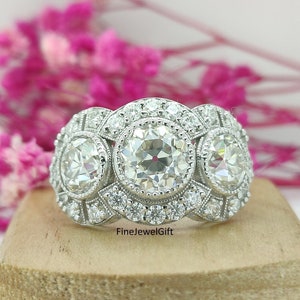 Puede incluir: Un anillo de plata con tres diamantes grandes redondos engastados en un diseño de halo. Los diamantes están rodeados de diamantes más pequeños. El anillo está sobre una superficie de madera.