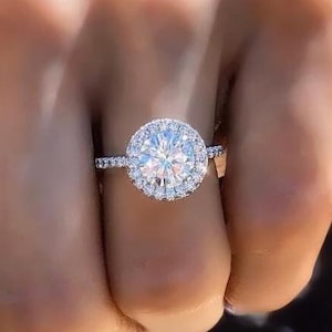 May include: A silver engagement ring with a round, clear center stone surrounded by a halo of smaller stones. The ring is set on a thin band.