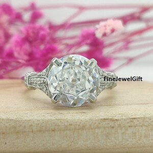 May include: A silver engagement ring with a large, round, clear gemstone surrounded by smaller gemstones. The ring has a delicate, intricate design with a band that tapers towards the center.