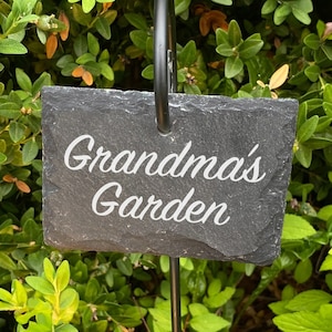 Könnte beinhalten: Ein rechteckiger Gartenmarker aus Schiefer mit den Worten "Grandma's Garden" in weißer Schrift. Der Marker ist an einem schwarzen Metallpfahl befestigt, inmitten von üppigem grünem Laub und rosa Blüten.