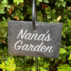 Könnte beinhalten: Ein schwarzer Gartenpfahl aus Metall mit einem Schieferschild, auf dem "Nana's Garden" steht. Der Pfahl ist von grünem Laub und rosa Blüten umgeben.