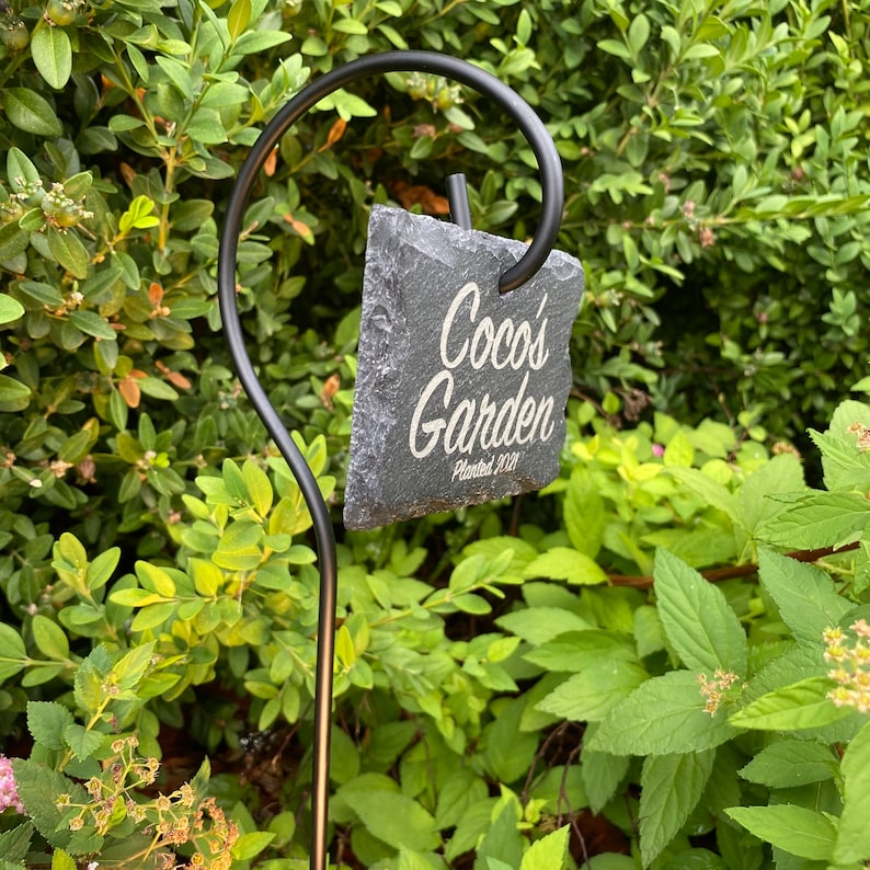 Slate garden sign 'Coco's Garden - Planted 2021' on a stake with dense greenery in the background.