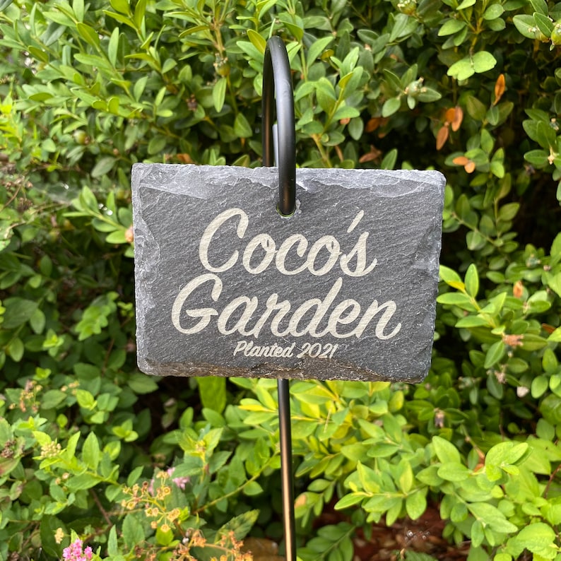 Custom engraved slate garden sign reading 'Coco's Garden' with the subtitle 'Planted 2021' hanging on a black metal stake in front of a lush green shrubbery background.